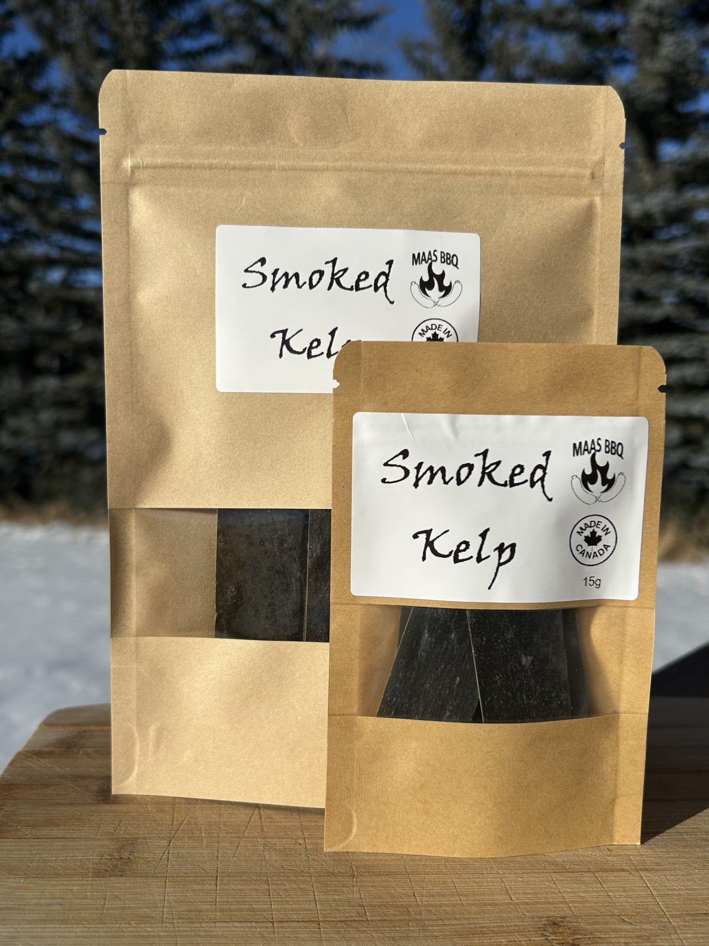 Two brown kraft pouches of Maas BBQ smoked kelp on a wooden surface, labeled “Smoked Kelp” with a “Made in Canada” stamp; culinary tags: vegan, gourmet, chef, BBQ, smoked kelp, Maas BBQ.
