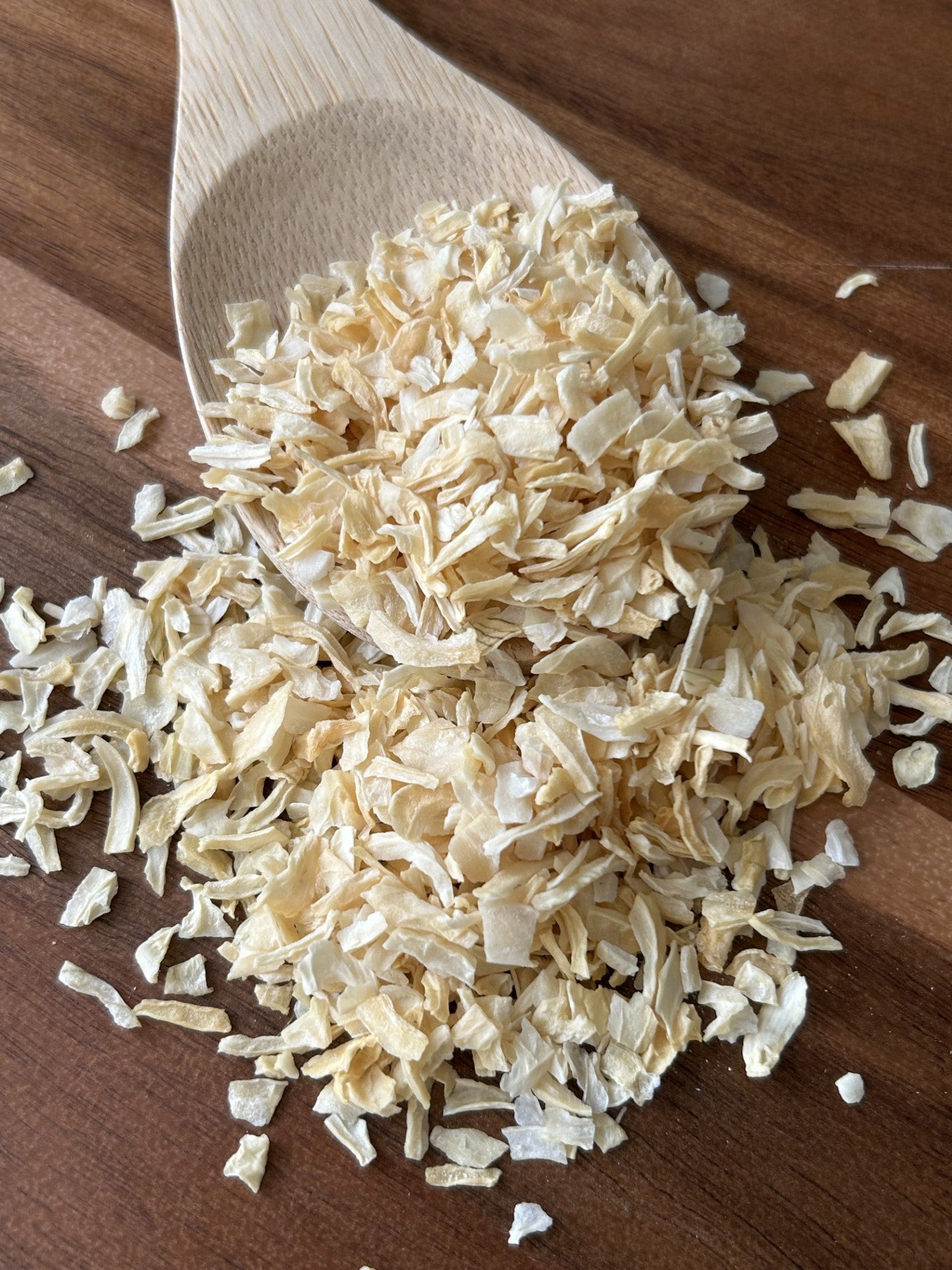 A wooden spoon rests on a brown wooden surface, holding a pile of light beige smoked onion flakes (vegan, plant‑based). Culinary tags: Maas BBQ, vegan, chef, gourmet, plant based, smoked onion flakes – use for seasoning BBQ dishes, soups, or vegan gourmet recipe