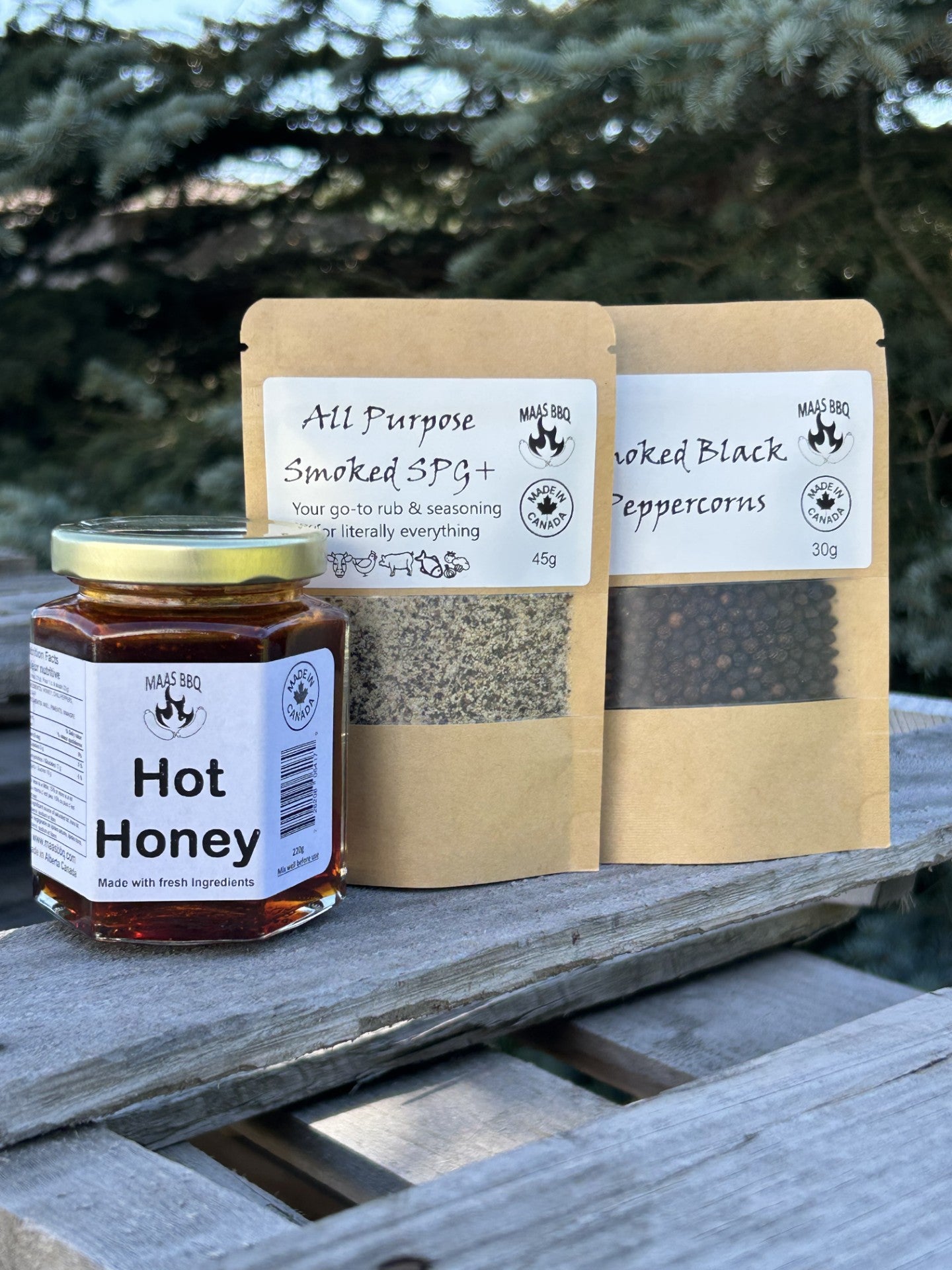 Jar of hot honey and bags of smoked SPG and black peppercorns on a wooden surface with a natural background.