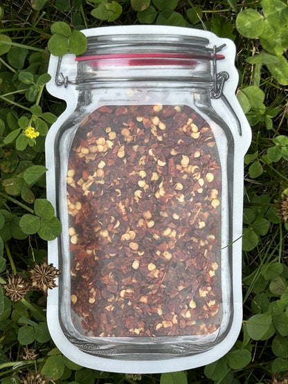 A unique mason jar‑shaped pouch of Maas BBQ Premium Smoked Red Pepper Flakes, showcasing the vibrant red flakes against a backdrop of green clover. This gourmet smoked spice, made in Alberta, Canada, is Canada's favorite for adding unique heat and smoky flavor to foodie cooking, BBQ, and diverse recipes, making it a great gift and pizza topping. 
