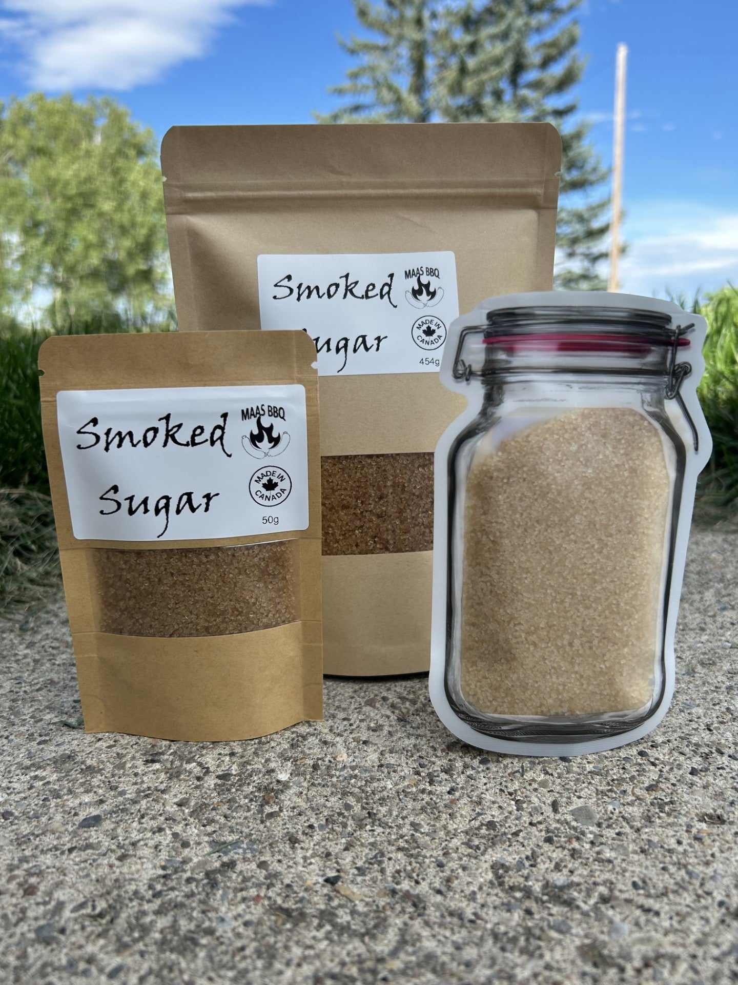 Maas BBQ Premium Smoked Sugar displayed in a 50g kraft pouch, a 454g kraft pouch, and a unique jar-shaped pouch, all against a natural outdoor background. This gourmet, unique smoked sugar is made in Alberta, Canada, perfect for adding distinct smoky-sweet flavor to cooking, BBQ recipes, desserts, and drinks. An ideal gift for any foodie or chef. Maas BBQ Premium Smoked Sugar – Canada’s Favorite Unique Gourmet Sweetener for Foodies & Chefs | Made in Alberta, Perfect for Cooking, BBQ & Gifts